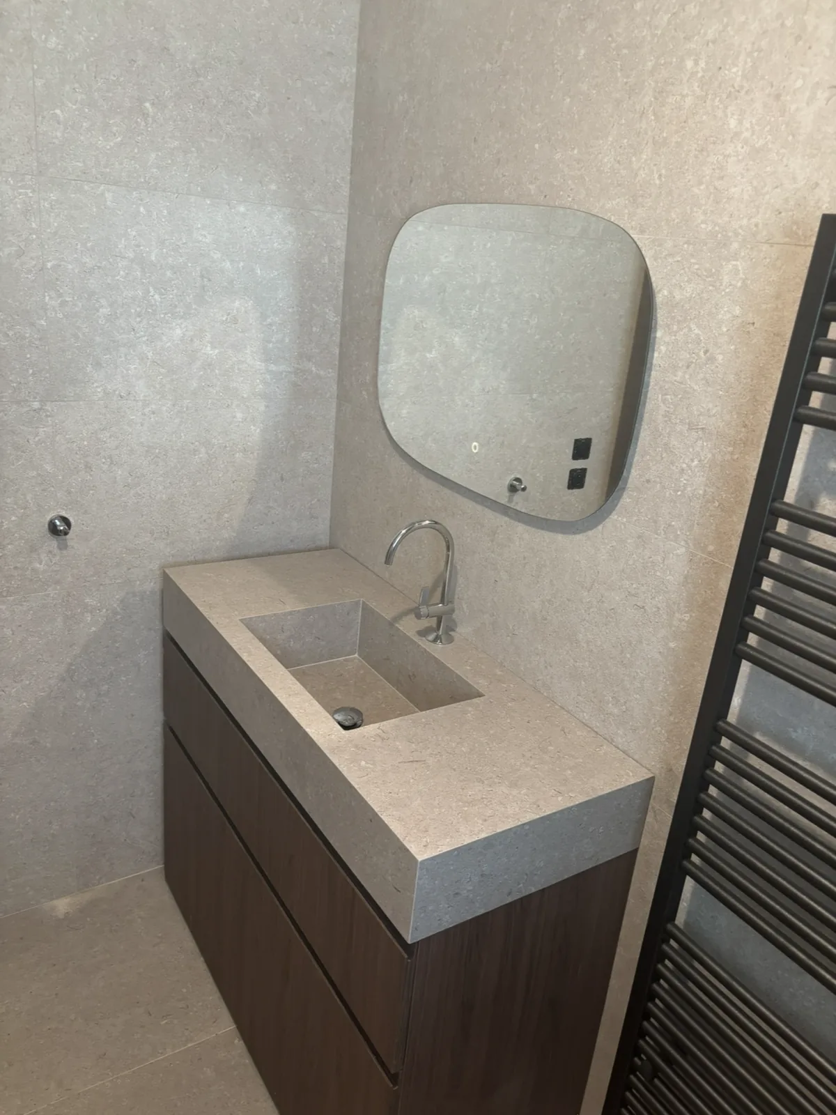Vasque sur mesure taillée dans le même carrelage effet pierre que les murs, posée sur un meuble en bois foncé, avec miroir lumineux.